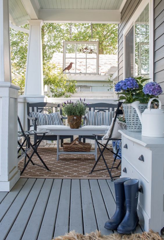 a vintage farmhouse summer porch with dark staiend furniture, striped pillows, rubber boots, a dresser and blooms