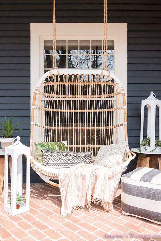 a welcoming summer porch with a large rattan hanging loveseat, an ottoman, large lanterns and potted greenery