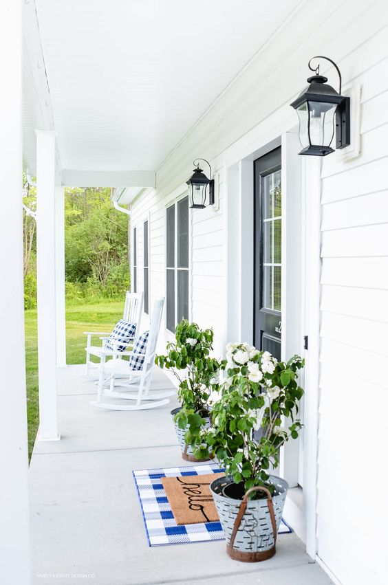 a white summer porch with comfy rocking chairs, potted greenery and blooms, a checked rug and lanterns