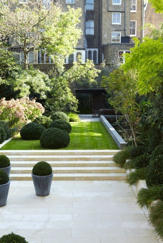 an elegant minimalist townhouse garden with stone tiles and steps, a lawn, boxwood, trees and shrubs plus a flower bed