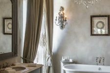 a French chic neutral bathroom with a clawfoot bathtub, a vanity, a mirror in an ornated frame, neutral textiles and a crystal chandelier