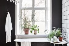 a black and white vintage bathroom with white square tiles, a black and white floor, a black statement wall, a crystal chandelier and lots of greenery
