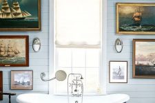 a chic and fancy bathroom with light blue planked walls, a navy tub and a fantastic marine gallery wall plus a glam gold lamp