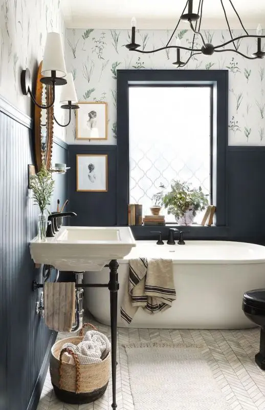 A contrasting bathroom with black paneled and floral print walls, a free standing bathtub and sink, a chandelier and artwork is pure elegance