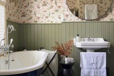 a cozy English country bathroom with floral wallpaper, green panels, a navy clawfoot bathtub, a free-standing sink and side tables