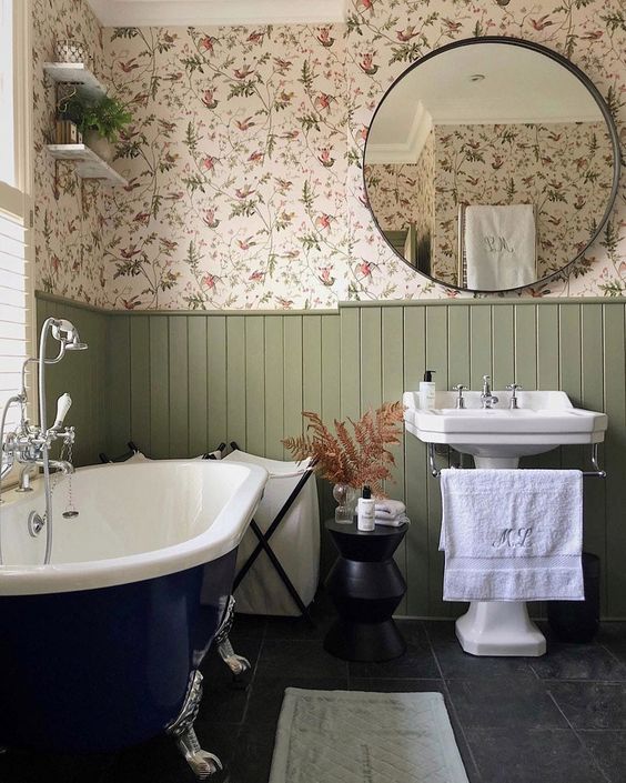 A cozy English country bathroom with floral wallpaper, green panels, a navy clawfoot bathtub, a free standing sink and side tables