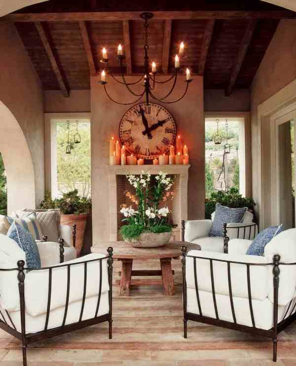 A cozy Mediterranean living room, outdoors and indoors, with a non working fireplace, a round wooden table, metal chairs with white upholstery and greenery