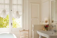 a delicate neutral bathroom with creamy paneling, a free-standing sink, a light-stained chair, a bathtub and a crystal chandelier