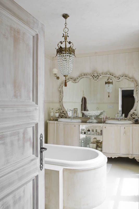 A jaw dropping vintage bathroom in neutrals, with a double vanity, a mirror in an ornated frame, a bathtub and a crystal chandelier