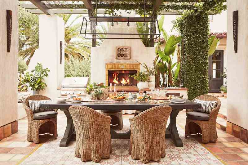 A refined Mediterranean outdoor dining space with greenery and potted plants, a dark stained table and wicker chairs, a pendant lamp and a fireplace