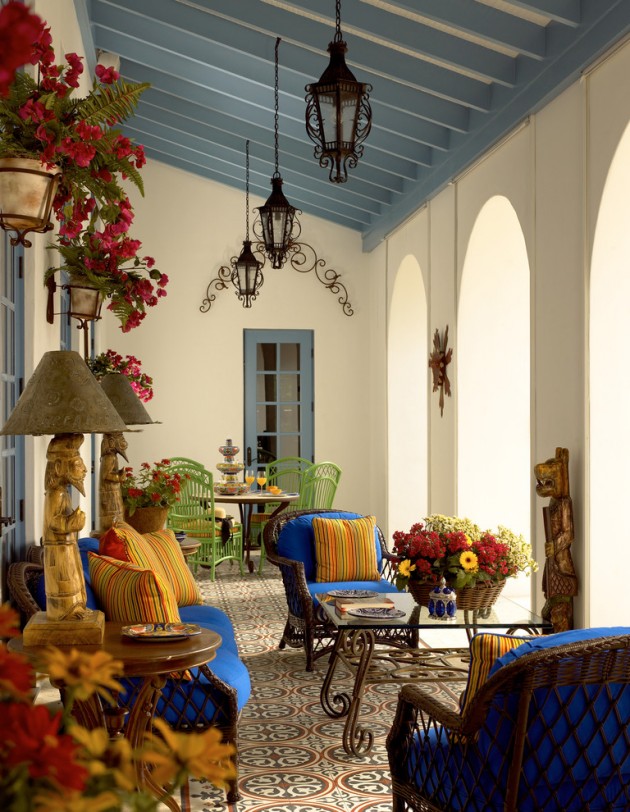 a refined Mediterranean patio done in bright colors, with a dining zone with a round table and green chairs, a glass table and bold blue wicker chairs, bright blooms and pendant lanterns