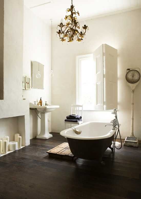A refined vintage bathroom with a faux fireplace, a black clawfoot bathtub, a free standing sink, a vintage chandelier and shutters on the window