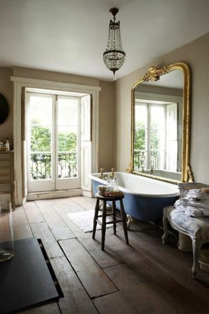 a vintage Parisian-inspired bathroom with tan walls, a blue clawfoot bathtub, an oversized mirror in a gilded frame, a crystal chandelier and vintage furniture
