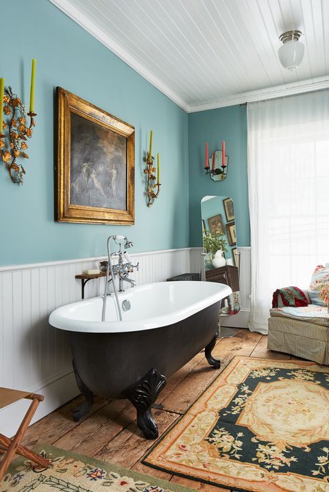 a vintage bathroom with blue walls, white paneling, a black clawfoot bathtub, artwork, printed rugs, a chair with printed pillows and colored candles