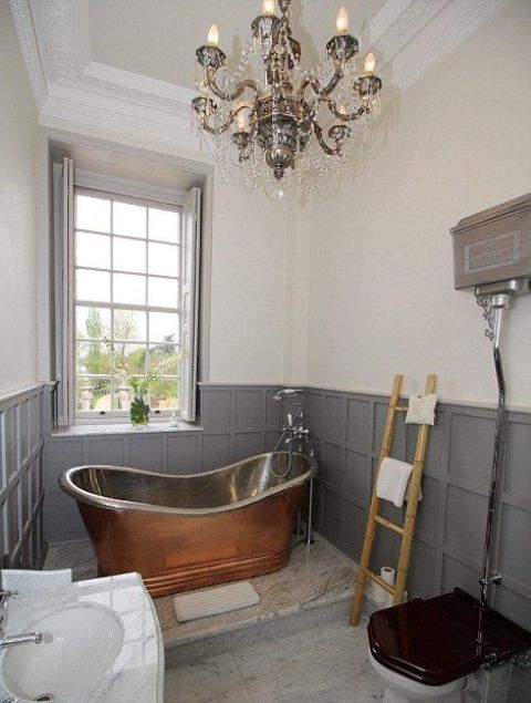 A vintage bathroom with grey paneling and shutters, a vintage metal bathtutb, a crystal chandelier, a fre standing sink and a ladder for towels