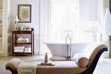 an airy vintage bathroom with a bathtub opposite the window, a dark-stained storage unit, a daybed with neutral upholstery and a pritned rug