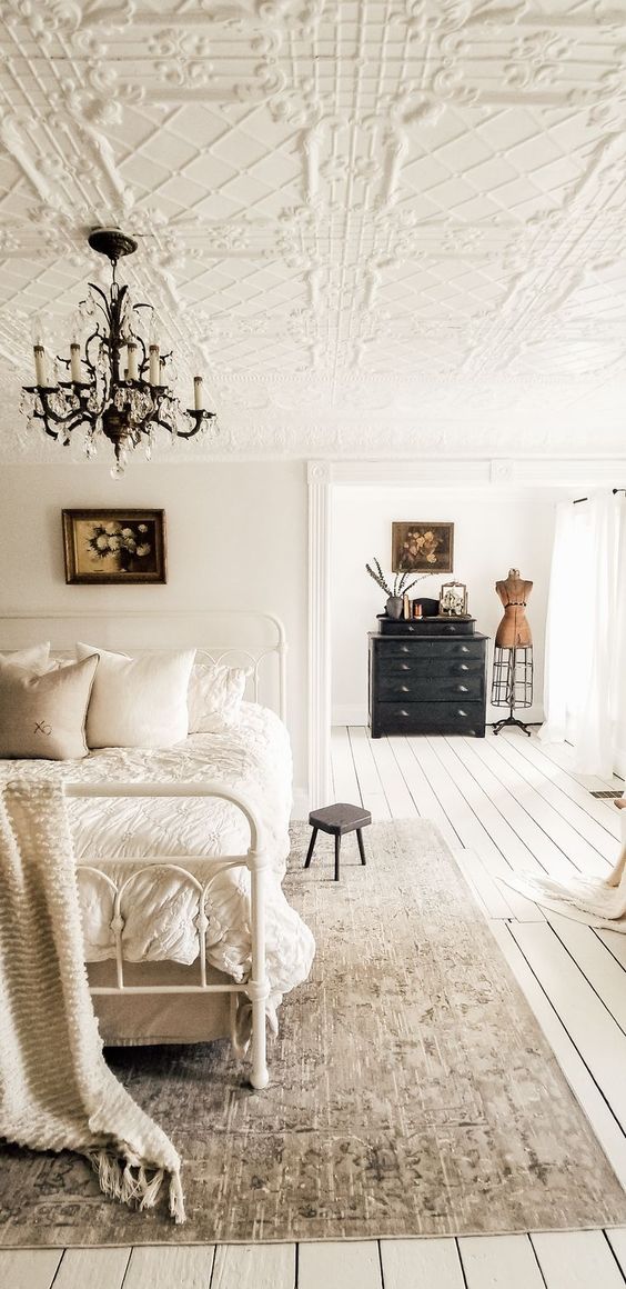 a beautiful Scandinavian bedroom with a chic vintage patterned ceiling, a white metal bed with neutral bedding, a black dresser and a lovely chandelier