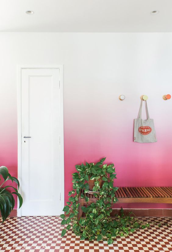 a bold entryway with a checked floor, an ombre white to pink wall, a stained bench and potted plants