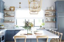 a chic coastal kitchen with ocean blue shaker cabinets, marble tiles, a large kitchen island with white countertops, wooden stools and gold pendant lamps