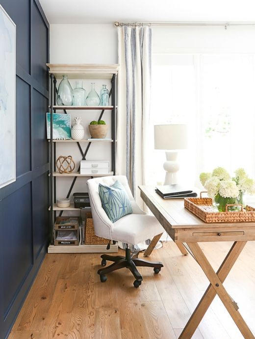 a coastal beach home office with a navy accent wall, a wooden trestle desk, an upholstered chair and a storage unit