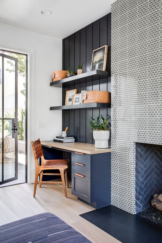 a coastal home office with a navy wall, a navy desk with a stone desktop, a wood and leather chair and a fireplace is welcoming