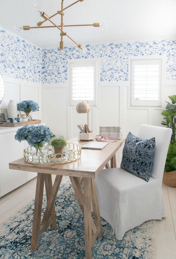 a coastal home office with blue printed wallpaper, white paneling, a trestle desk, an upholstered chair and a gilded chandelier