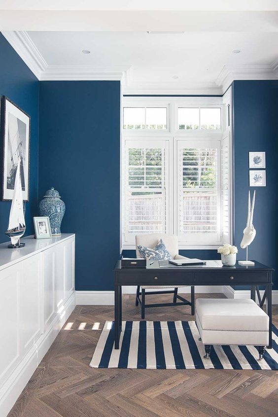 a coastal home office with navy walls, a white storage unit, a navy desk, a white ottoman and cool sea artworks