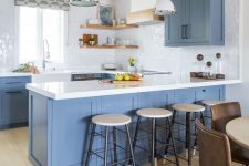 a fab coastal kitchen with blue cabinets, white stone countertops and glossy tiles, light-stained shelves and wooden beams