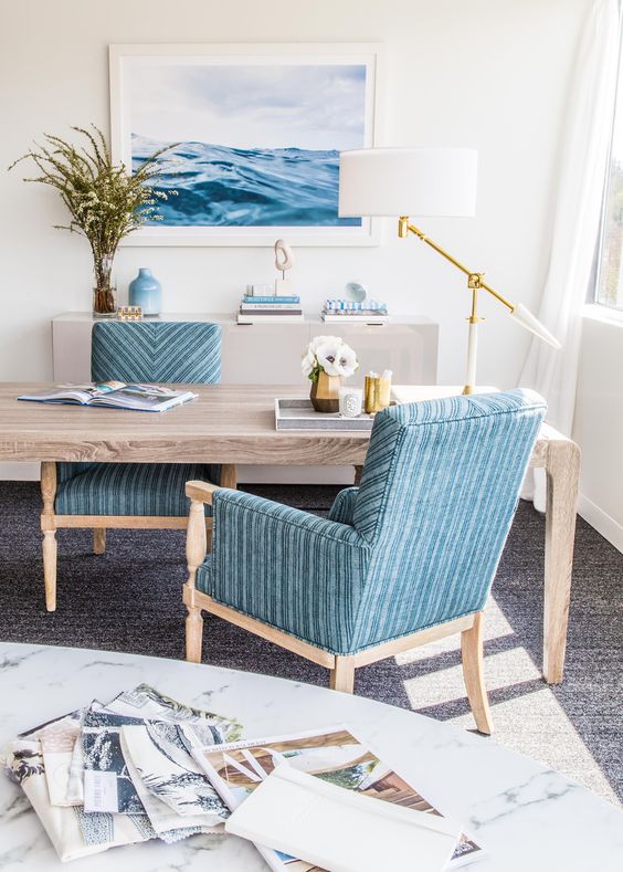 a gorgeous seaside home office with a wooden desk, blue chairs, a sea-inspired artwork, an elegant and catchy table lamp
