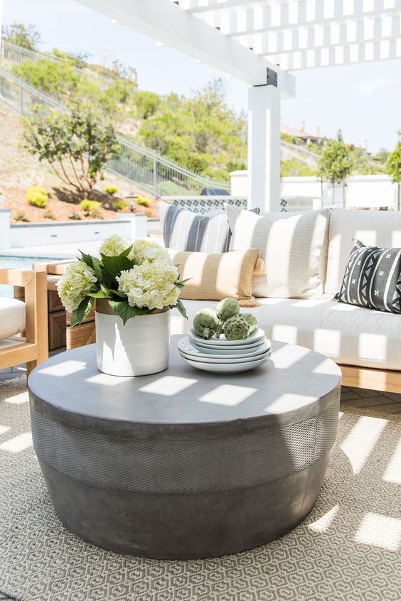A light filled terrace with a neutral sofa with printed pillows, a chair, a round concrete coffee table and some blooms
