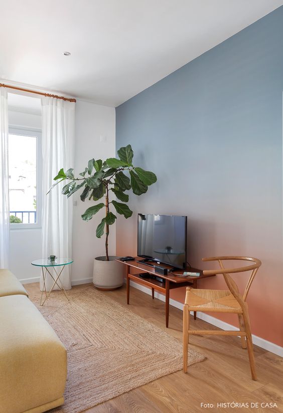 A living room with a gradient blue and red wall, a yellow sofa, a TV on a stand, a mid century modern chair and a tree in a pot