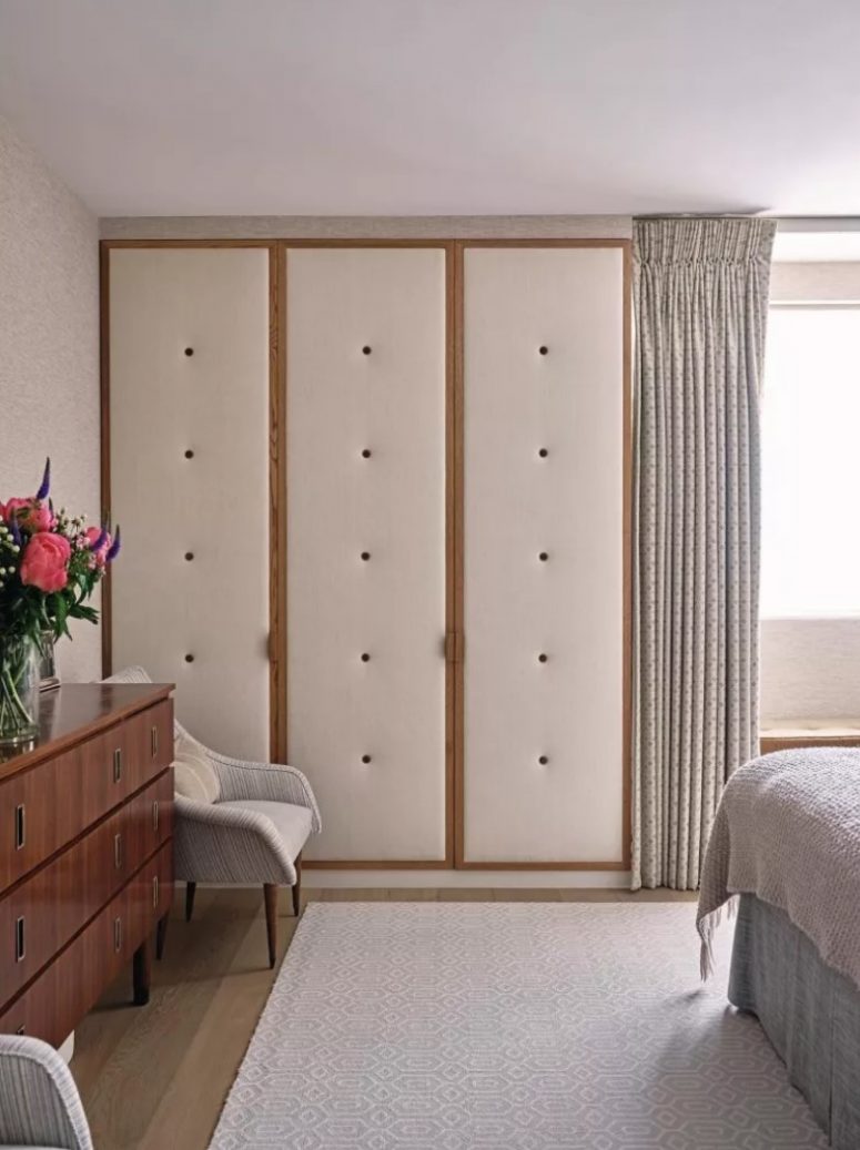 a modern and chic bedroom done in neutrals, with a bed with neutral bedding, a stained dresser, a wardrobe with uphlostered panels and wooden frames