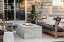 a modern outdoor space with wooden sofas with lots of pillows, a concrete fireplace, woven pendant lamps and a cocnrete stool