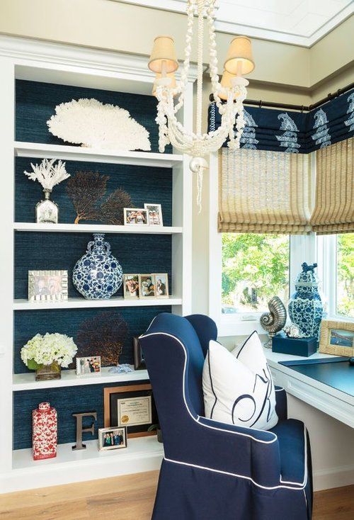a nautical home office with navy walls, a white storage unit, a white desk, a navy chair, a chic chandelier and shades and curtains