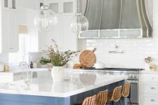 a pretty and bright coastal kitchen with white shaker style cabinets, a blue kitchen island, glass sphere pendant lamps and woven stools