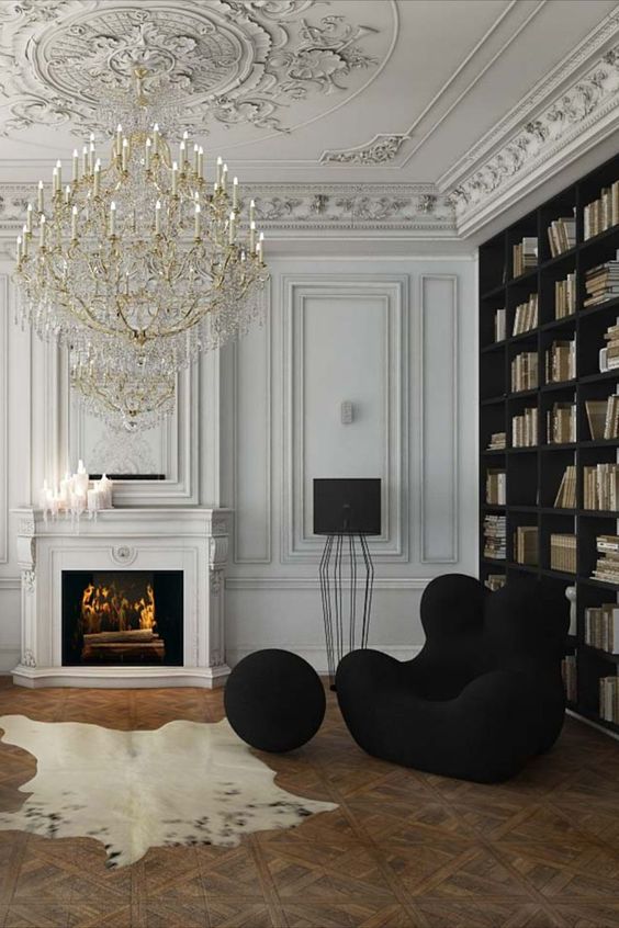 A sophisticated Parisian living room with a stucco ceiling, paneled walls, a built in bookcase, a fireplace, a black chair and a pouf and an oversized chandelier