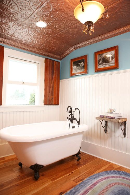 A vintage bathroom with white paneling and blue walls, copper tin tiles on the ceiling, a clawfoot bathtub, a wall mmounted shelf, artwork