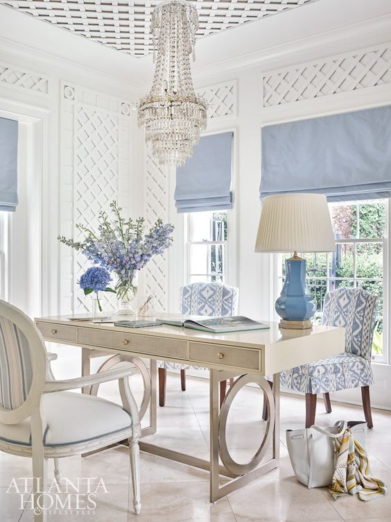 a vintage coastal home office with paneling on the walls, a neutral desk, chic printed chairs, a crystal chandelier and blue curtains