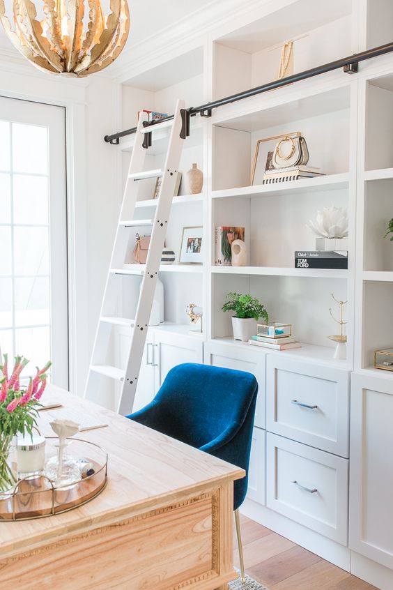 A white coastal home office with a large storage unit with a ladder, a light stained desk and a navy chair plus a catchy chandelier