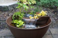 an aqua garden of a large bowl with a pond with pebbles and plants, a fountain and blooms and greenery