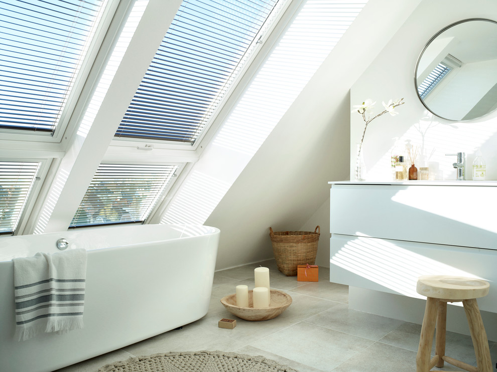 a contemporary attic bathroom with large windows, an oval bathtub, a vanity with a round mirror, a wooden stool  (VELUX)