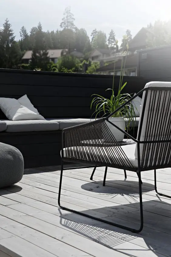A Nordic terrace with a grey deck, a black built in bench, a black chair with white upholstery and greenery