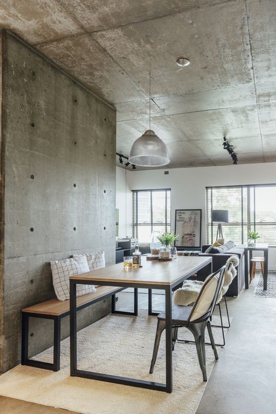 a contemporary industrial dining space with rough concrete walls and a ceiling, a minimal wood and metal table, a matchign bench and metal chairs