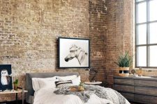 a light-flooded industrial bedroom with brick walls, a reclaimed wood dresser, a bench, a grey upholstered bed with neutral bedding, exposed piping