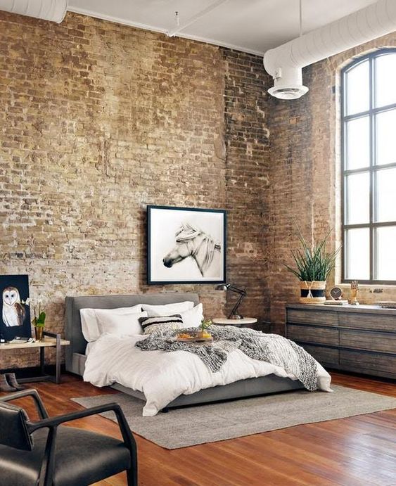 A light flooded industrial bedroom with brick walls, a reclaimed wood dresser, a bench, a grey upholstered bed with neutral bedding, exposed piping