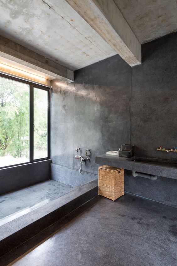 A minimalist industrial bathroom done with white concrete and black stone, a large built in tub, a vanity with a built in sink and a basket