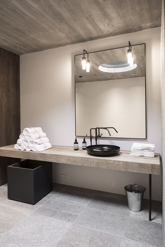 a minimalist industrial bathroom with stone tiles, concrete walls, a rough wooden vanity, a large mirror and lamps