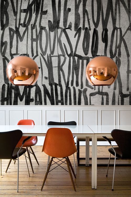 a modern dining room with a graffiti all over the wall, neutral paneling, white tables, black and orange chairs and copper sphere lamps