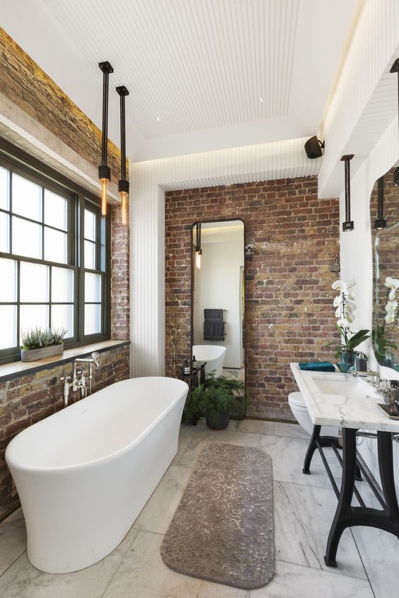A modern industrial bathroom with brick walls, a stone sink on a stand, a free standing tub, exposed pipes and bulbs
