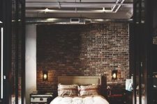 a refined industrial bedroom with brick walls, an upholstered bed, a chest and a dresser and exposed pipes on the ceiling